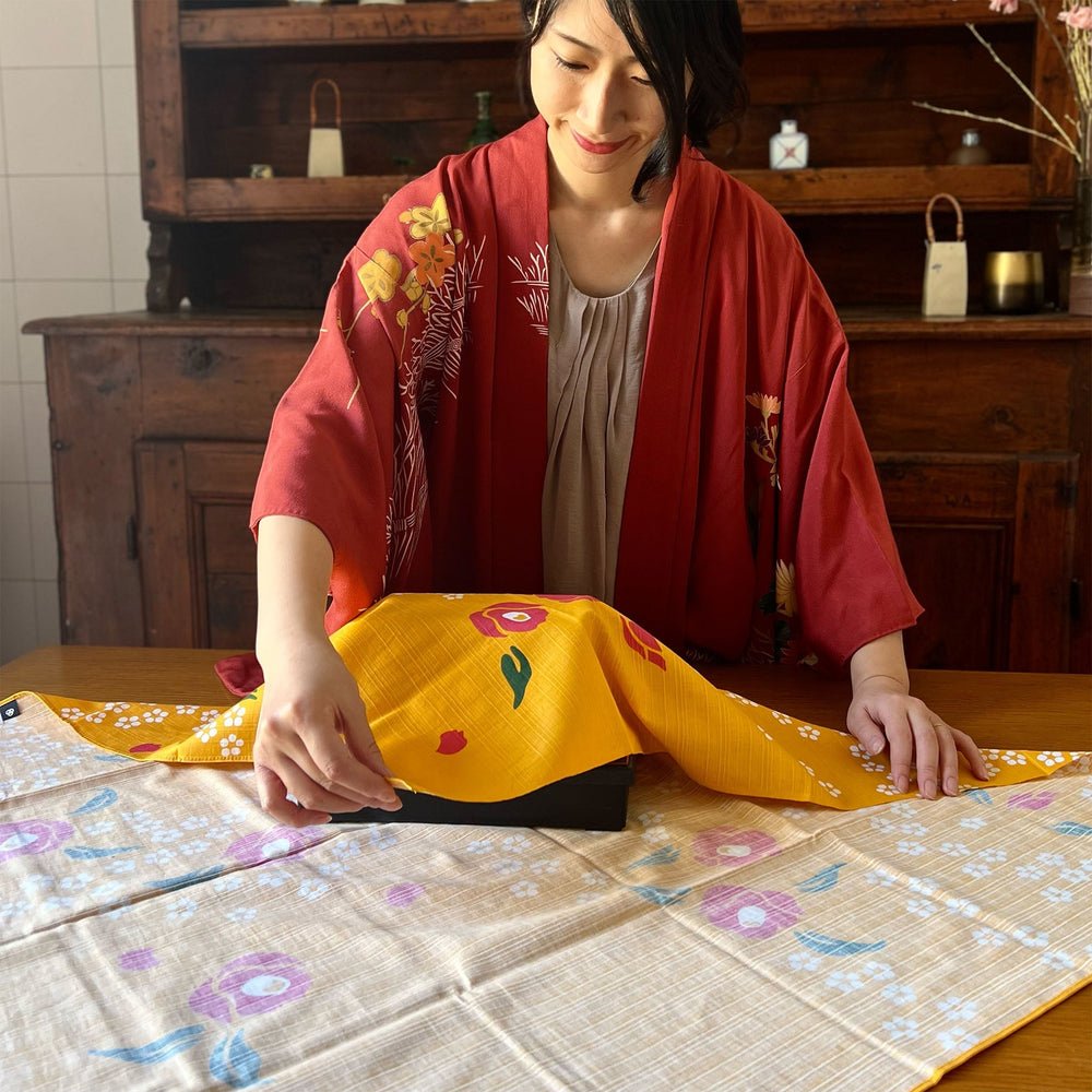 
                  
                    Furoshiki Camelia e Spirea - Kenema (90 x 90 cm) | Todoku Japan. Megumi utilizza Furoshiki per avvolgere una scatola.
                  
                